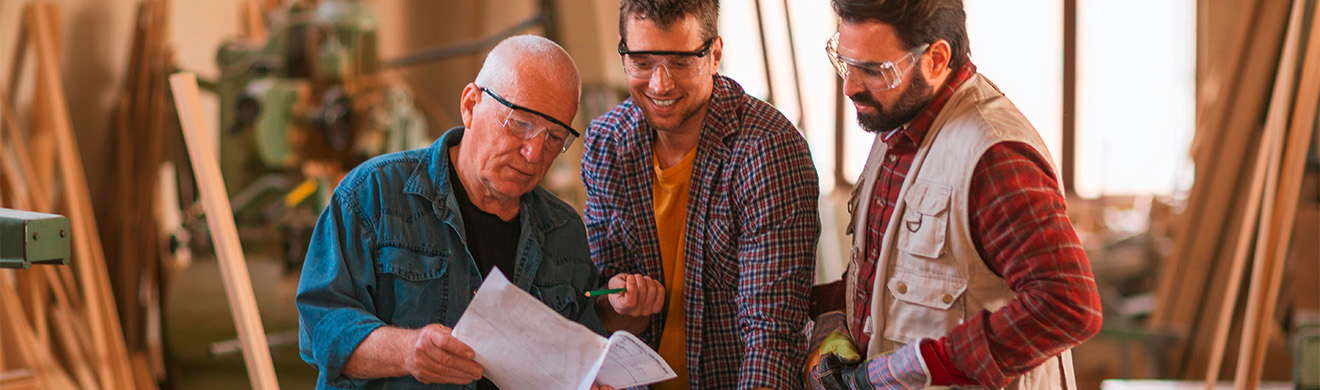 Mehrere Tischler verschiedener Generationen arbeiten gemeinsam in einer Holzwerkstatt an individuellen Möbel- und Innenausbauprojekten.